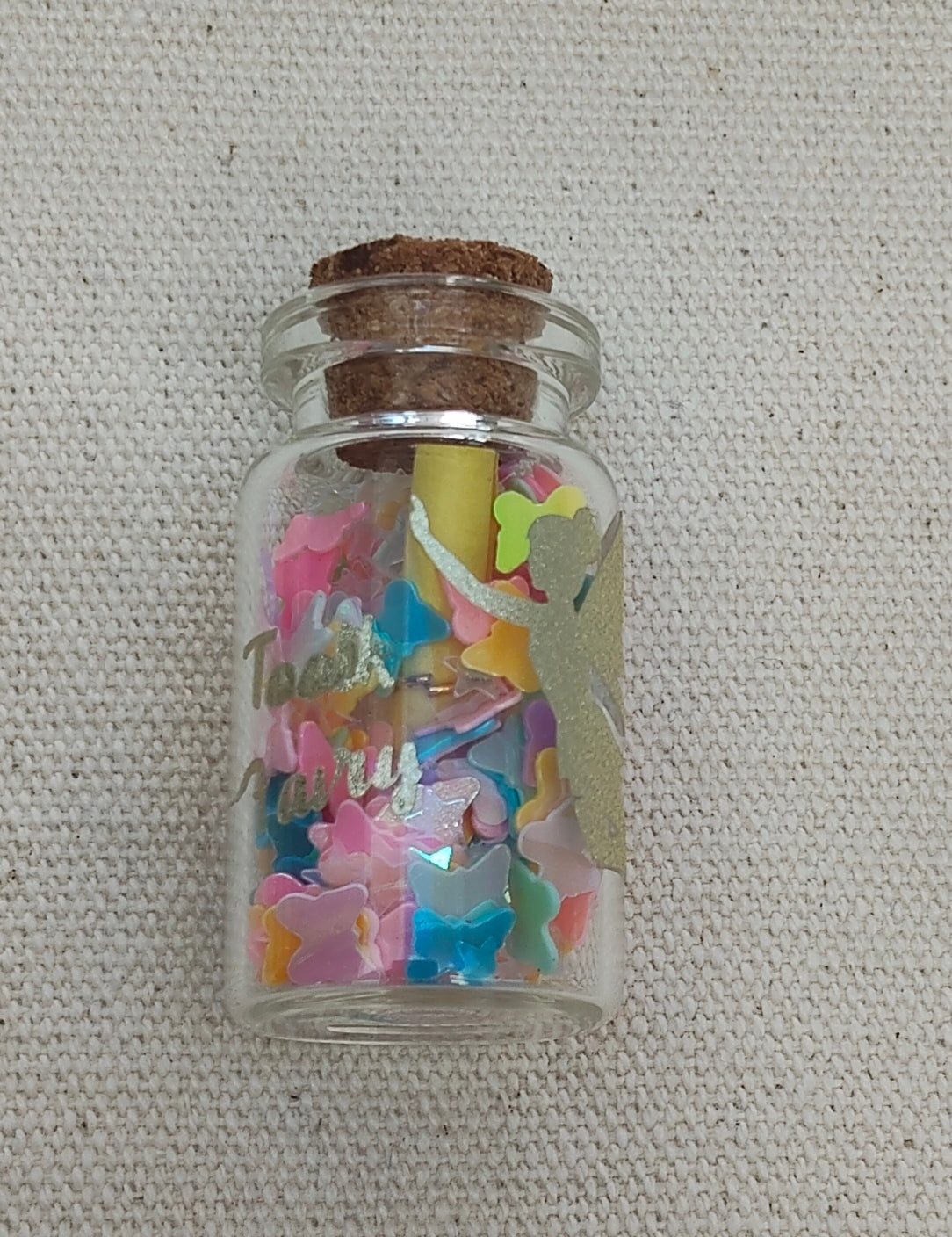 Small glass jar with colorful butterfly-shaped confetti and a cork lid, with tooth fairy design on the glass, with a message in the bottle from the Tooth Fairy. 